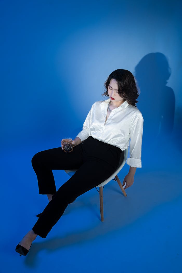 Fashionable woman in formal attire relaxing with a drink in a blue-lit studio.